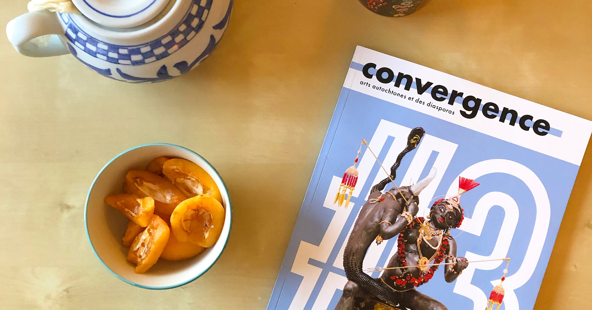 Third issue of Convergence, a blue cover with a sculpture of by Jaishri Abichandani, on a table with tea and apricots